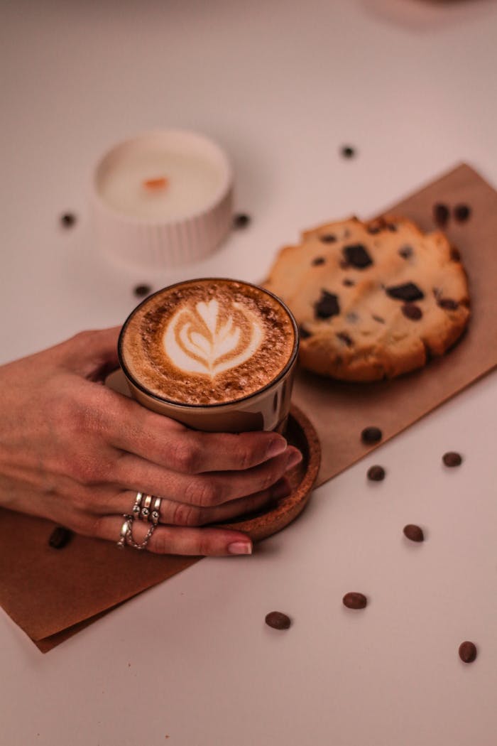 why-choose-us Aesthetic flat lay of cappuccino with latte art, chocolate chip cookie, and a candle on a wooden table.